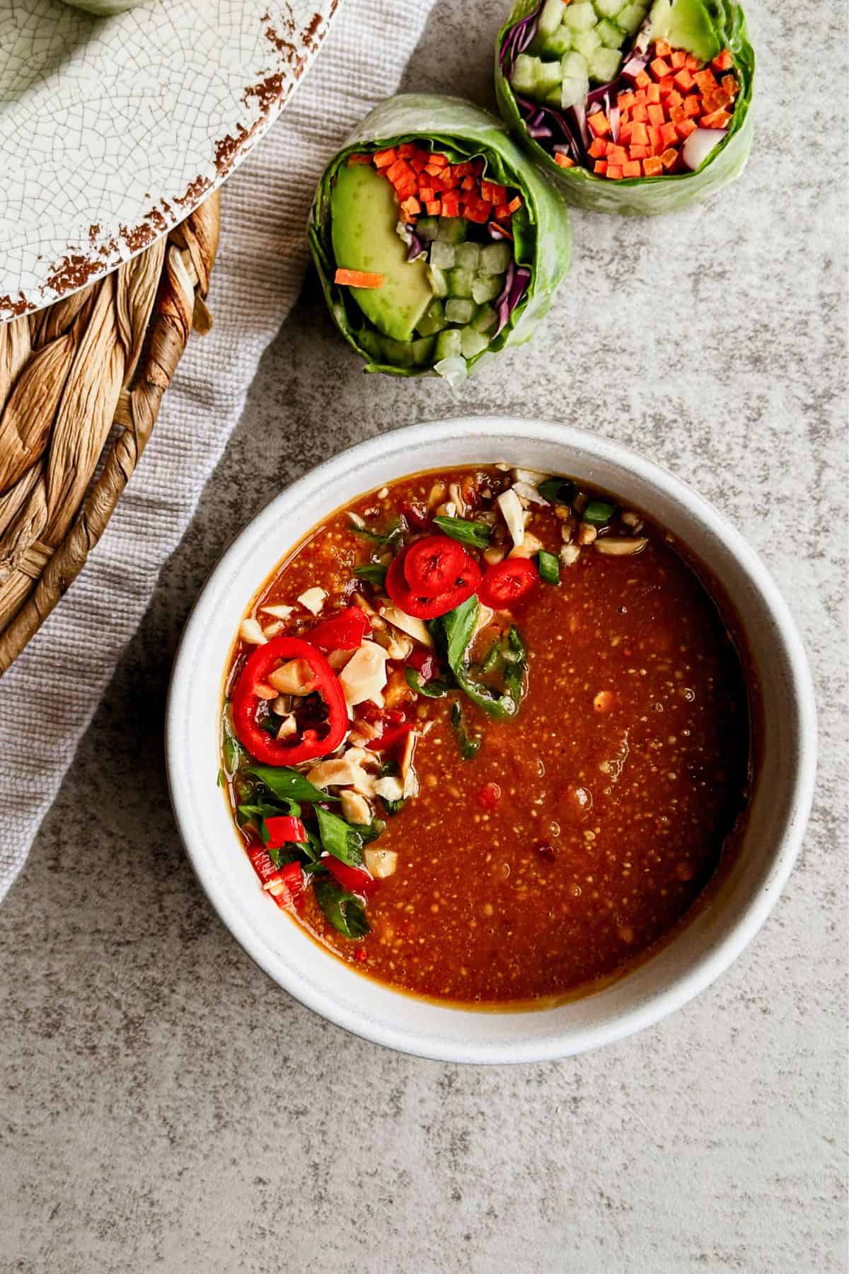 Bowl of peanut sauce garnished with sliced red chili and peanuts, served with fresh spring rolls.