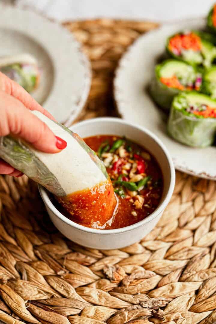 Hand dipping a fresh vegetable spring roll into a bowl of spicy Thai peanut sauce, garnished with peanuts and chili.