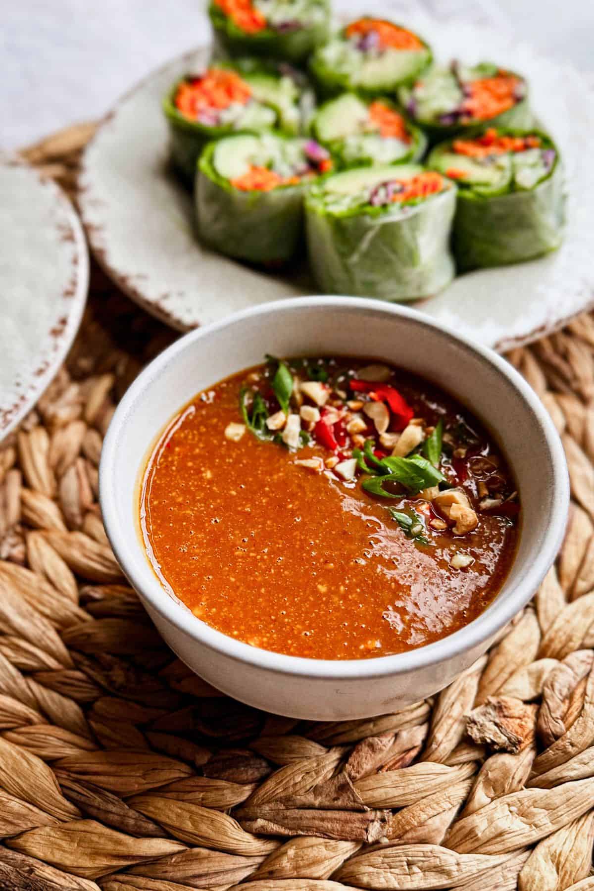 Peanut sauce in a bowl, garnished with chopped peanuts, scallions, and red chili, served with fresh vegetable spring rolls