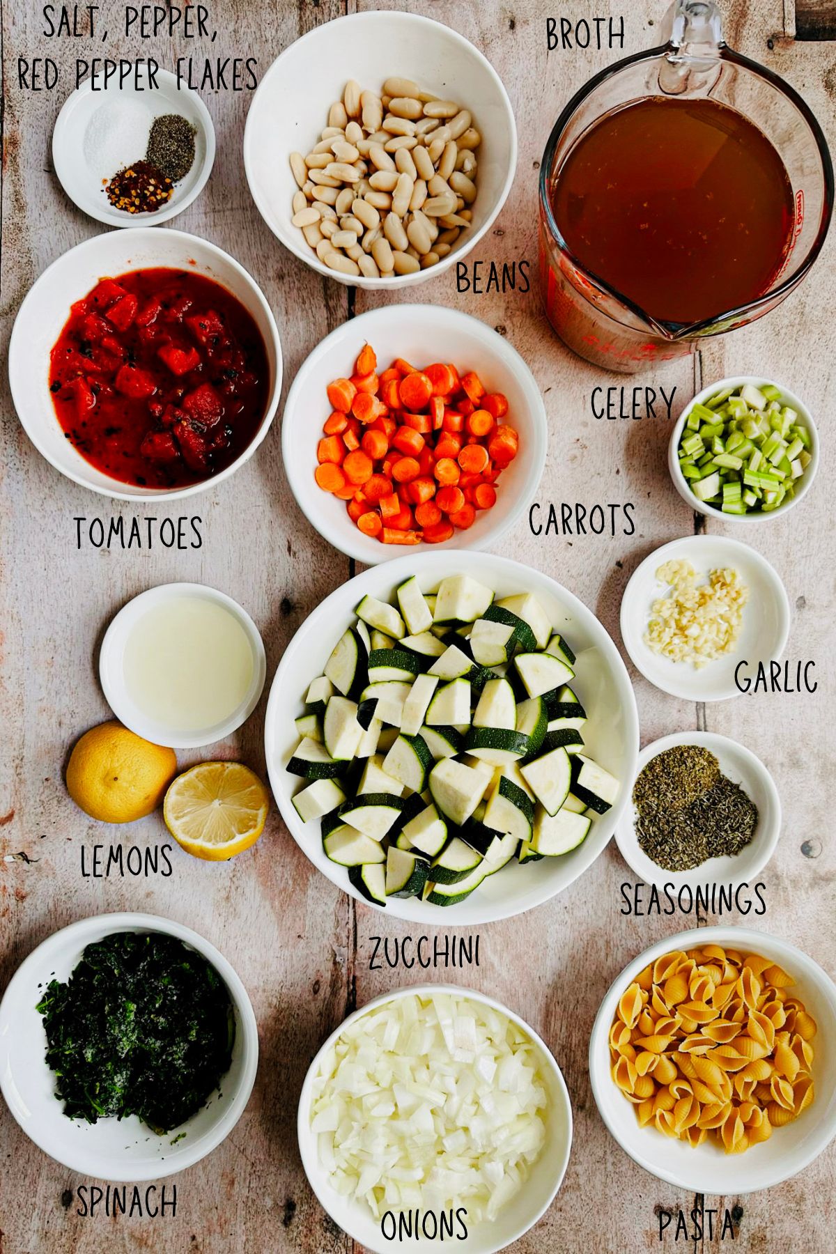 Ingredients for vegan minestrone soup including cannellini beans, zucchini, spinach, carrots, celery, onions, garlic, tomatoes, pasta, broth, lemons, and seasonings in white bowls on a wooden surface.