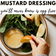 A hand dipping an apple slice into a jar of creamy vegan honey mustard dressing nestled in a bowl of fresh spinach and apple slices, with text reading 'How to Make Vegan Honey Mustard Dressing you'll never know is egg free.'