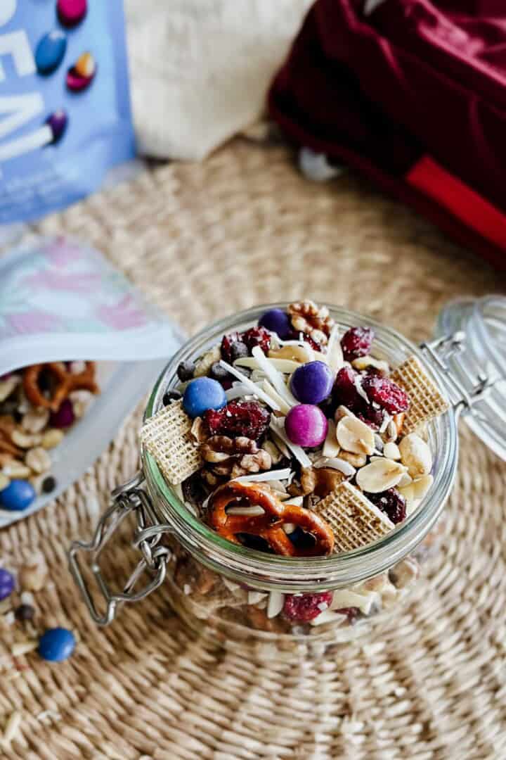 Trail Mix Glass jar filled with colorful vegan trail mix including pretzels, peanuts, walnuts, dried cranberries, slivered almonds, coconut shreds, whole grain cereal squares, chocolate chips, and Real Gems chocolate candies, with a woven mat background and snack bags nearby.