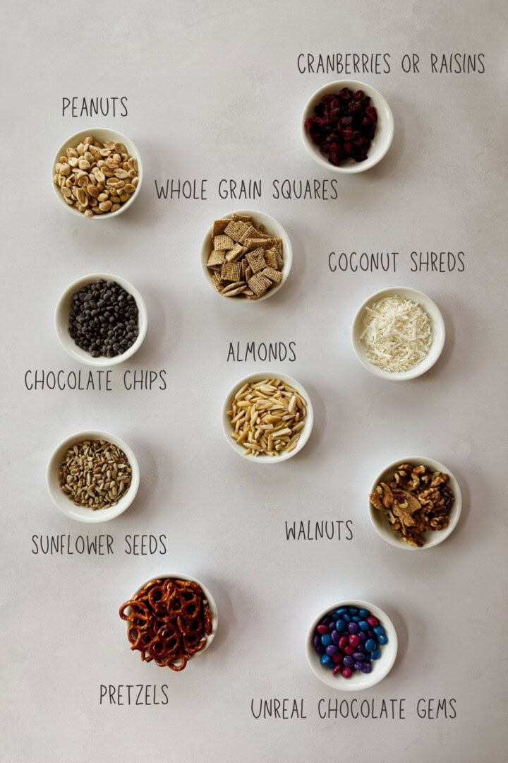 Trail mix ingredients Overhead image of small white bowls filled with individual vegan trail mix ingredients, including peanuts, chocolate chips, sunflower seeds, whole grain cereal squares, cranberries, coconut shreds, almonds, walnuts, pretzels, and colorful Real Gems chocolate candies. Each bowl is labeled with simple black text on a light background.