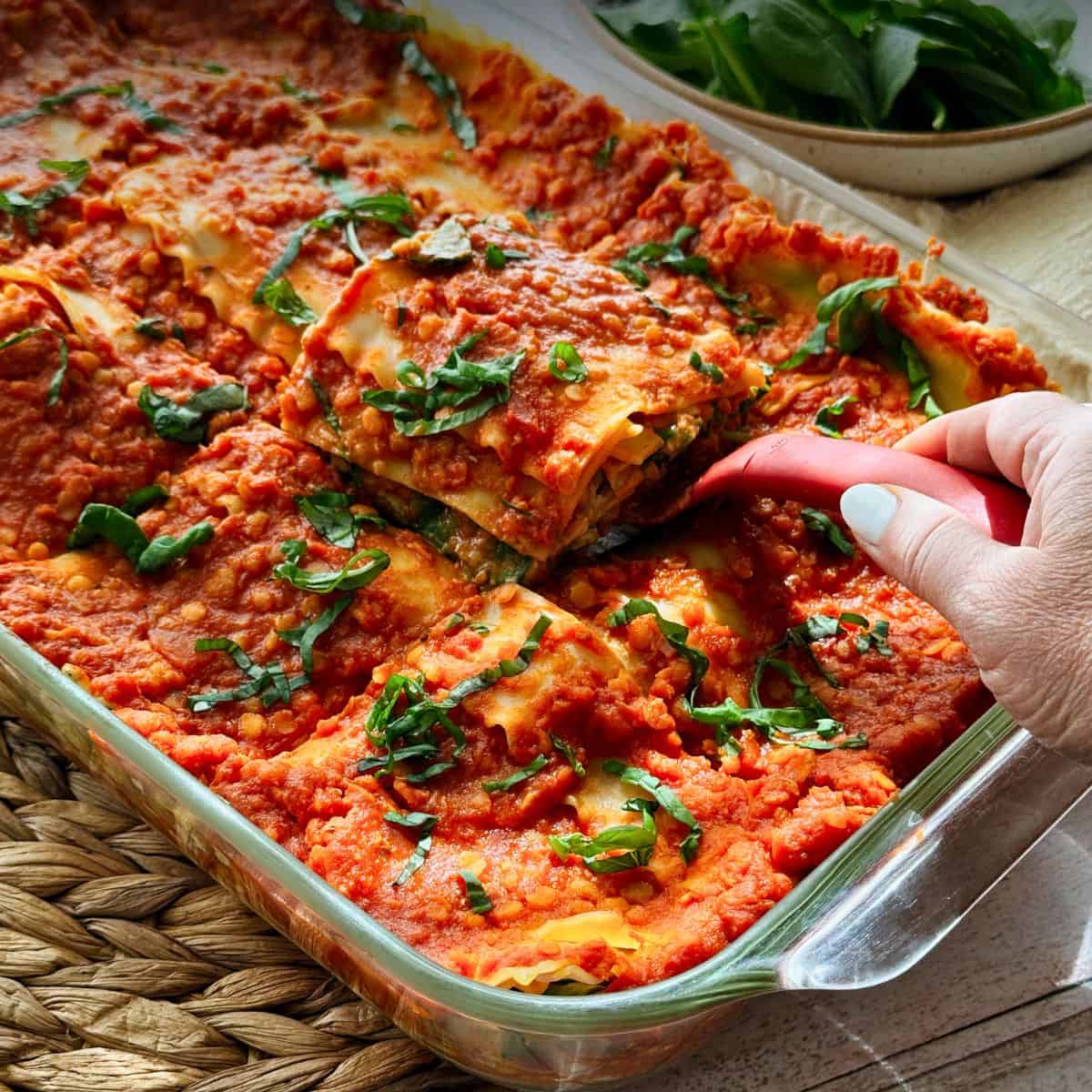 Close-up of vegan lasagna with marinara and basil. A hand lifts a slice, showing layers of noodles, spinach, and creamy filling.