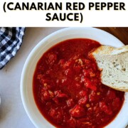 A white ceramic bowl filled with chunky Canarian red pepper sauce (mojo picón), deep red and glossy with visible roasted pepper pieces, with a thick slice of crusty sourdough bread dipping into it.