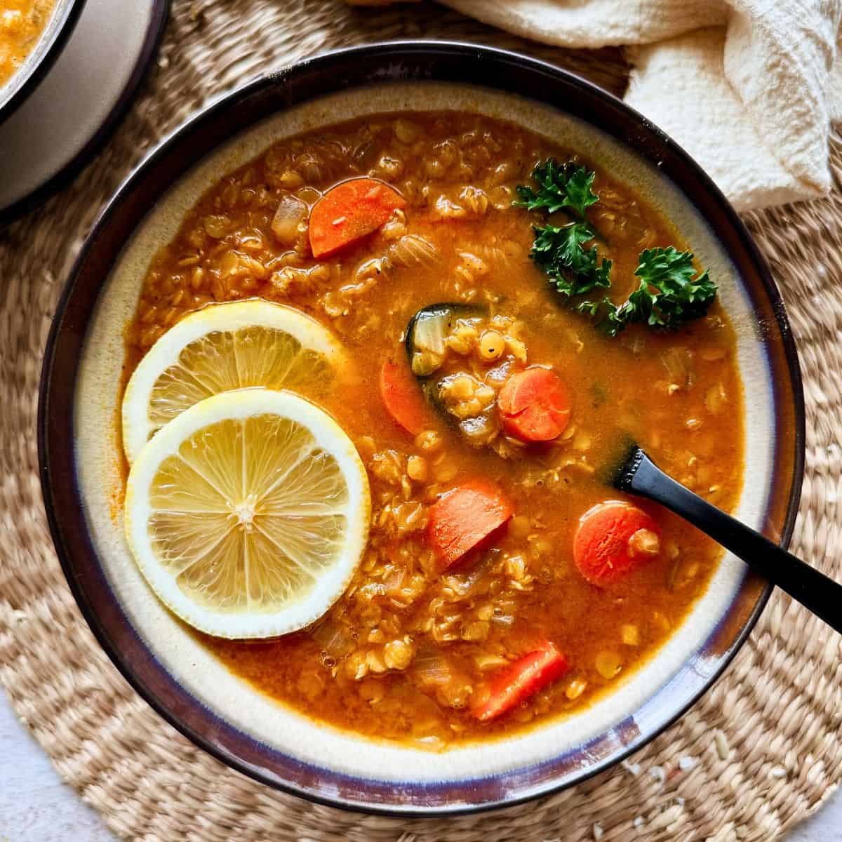A bowl of red lentil soup is garnished with lemon slices and fresh parsley, filled with tender carrots and a rich, golden broth. A black spoon rests in the bowl, ready to serve.