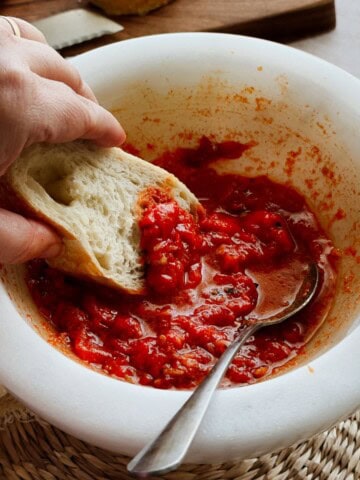 Hand dipping a piece of rustic bread into a bowl of vibrant red mojo picón sauce, with a spoon resting in the bowl.