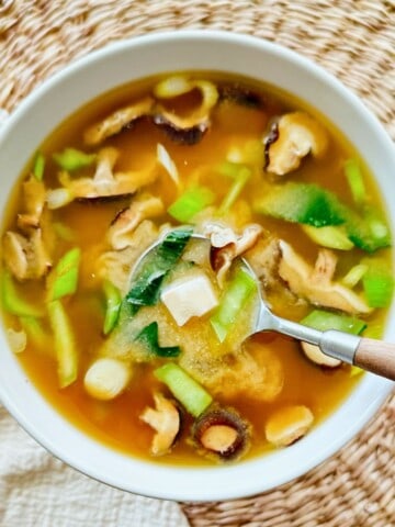 Bowl of vegan miso soup filled with shiitake mushrooms, bok choy, scallions, and silken tofu cubes. A spoon rests inside the bowl, lifting broth and vegetables, set on a woven placemat with a light cloth napkin.