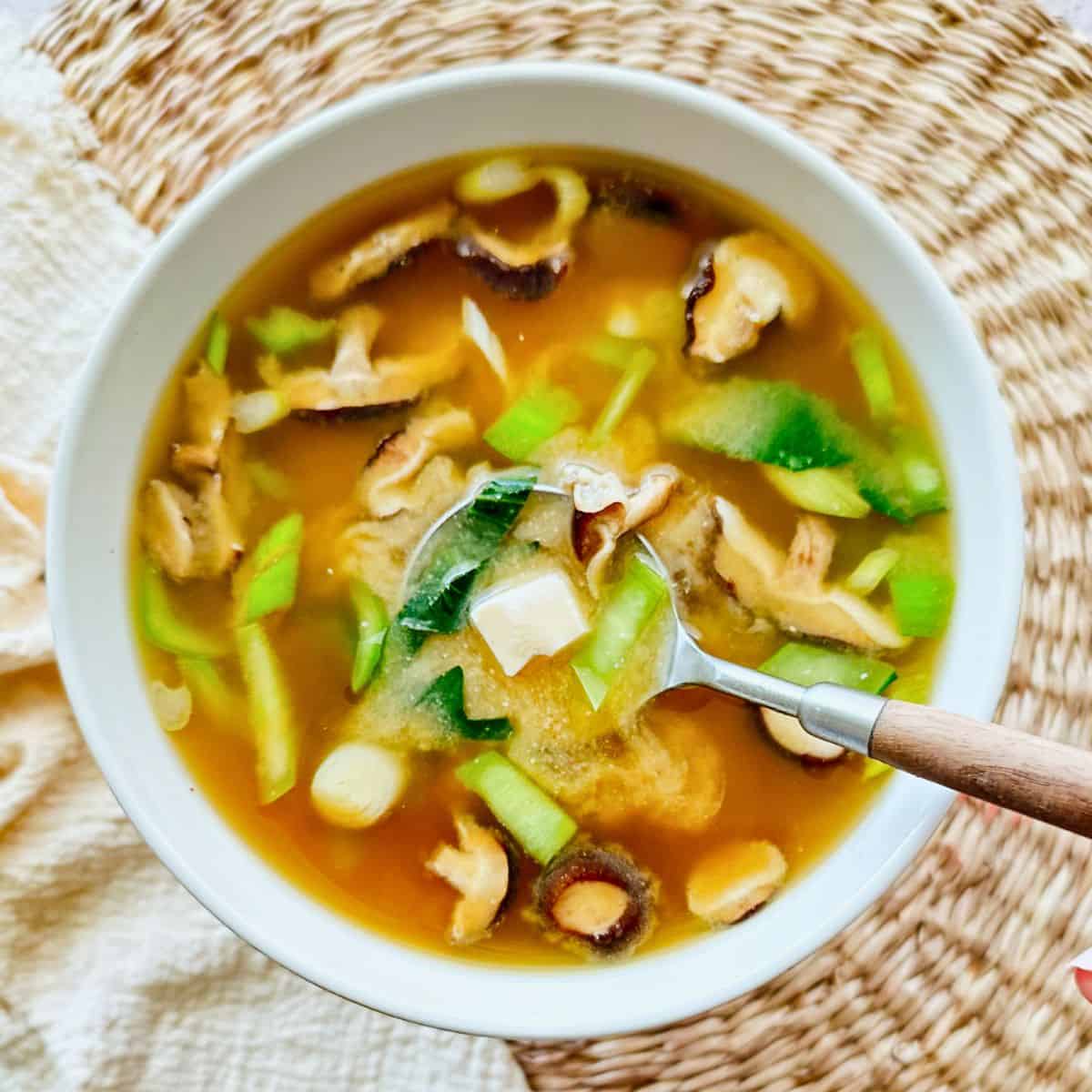 Bowl of vegan miso soup filled with shiitake mushrooms, bok choy, scallions, and silken tofu cubes. A spoon rests inside the bowl, lifting broth and vegetables, set on a woven placemat with a light cloth napkin.