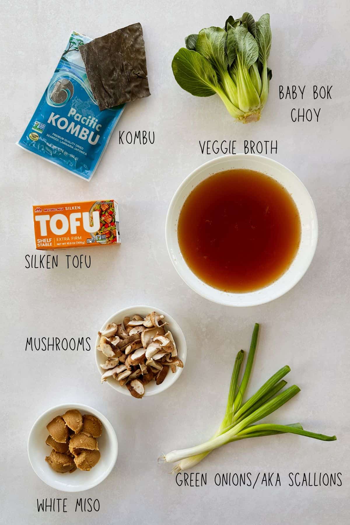 Flat lay of vegan miso soup ingredients on a light surface, each labeled with text: a package of kombu seaweed, a bunch of baby bok choy, a bowl of veggie broth, a box of silken tofu, a small bowl of sliced mushrooms, a small bowl of white miso paste, and fresh green onions also known as scallions.