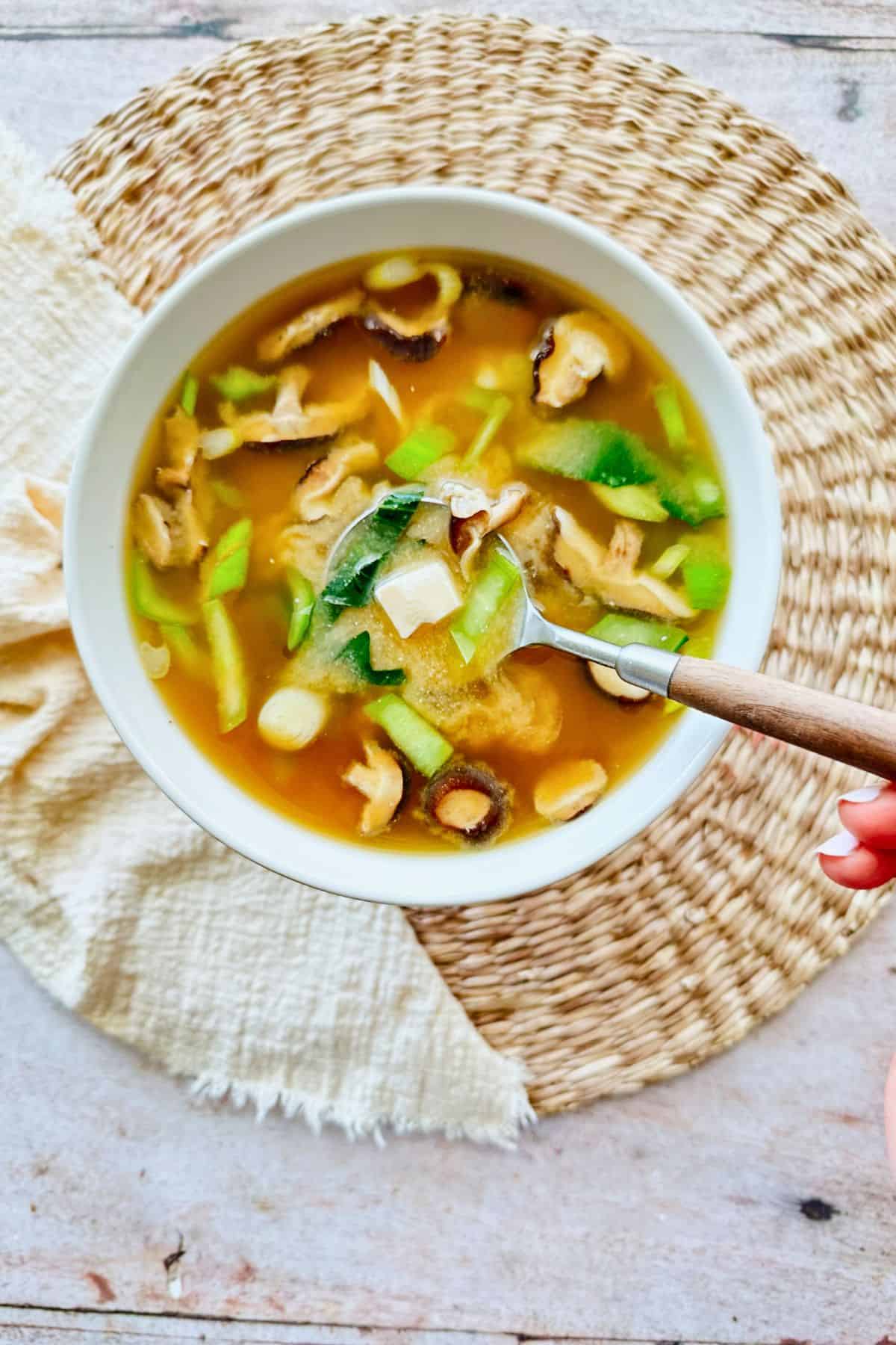 Bowl of vegan miso soup with mushrooms, bok choy, scallions, and tofu cubes in a light brown broth. A spoon is lifting a piece of tofu and vegetables from the bowl, set on a woven placemat with a cream cloth napkin.