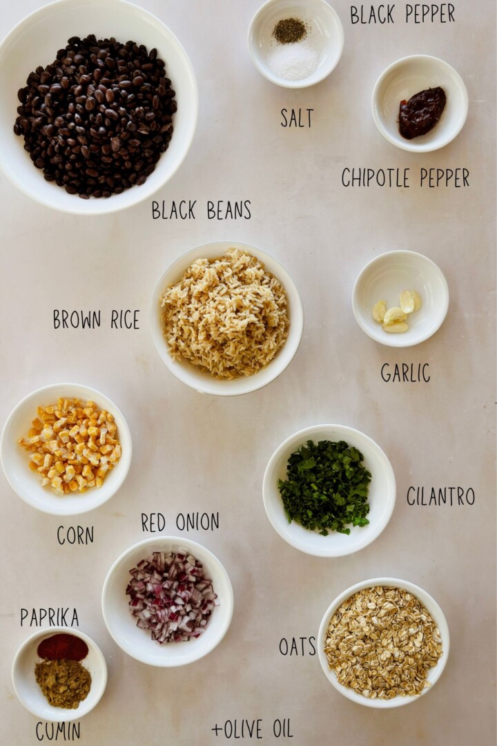 Overhead view of labeled chipotle black bean burger ingredients arranged in bowls, including black beans, brown rice, oats, corn, red onion, garlic, cilantro, chipotle pepper, paprika, cumin, salt, black pepper, and olive oil.