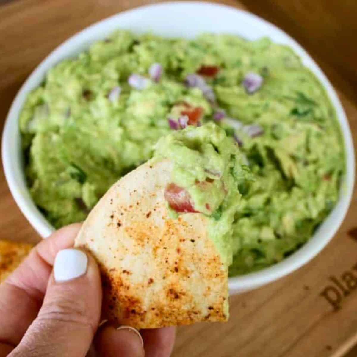 chip held by a hand with Guacamole on it; bowl is behind it
