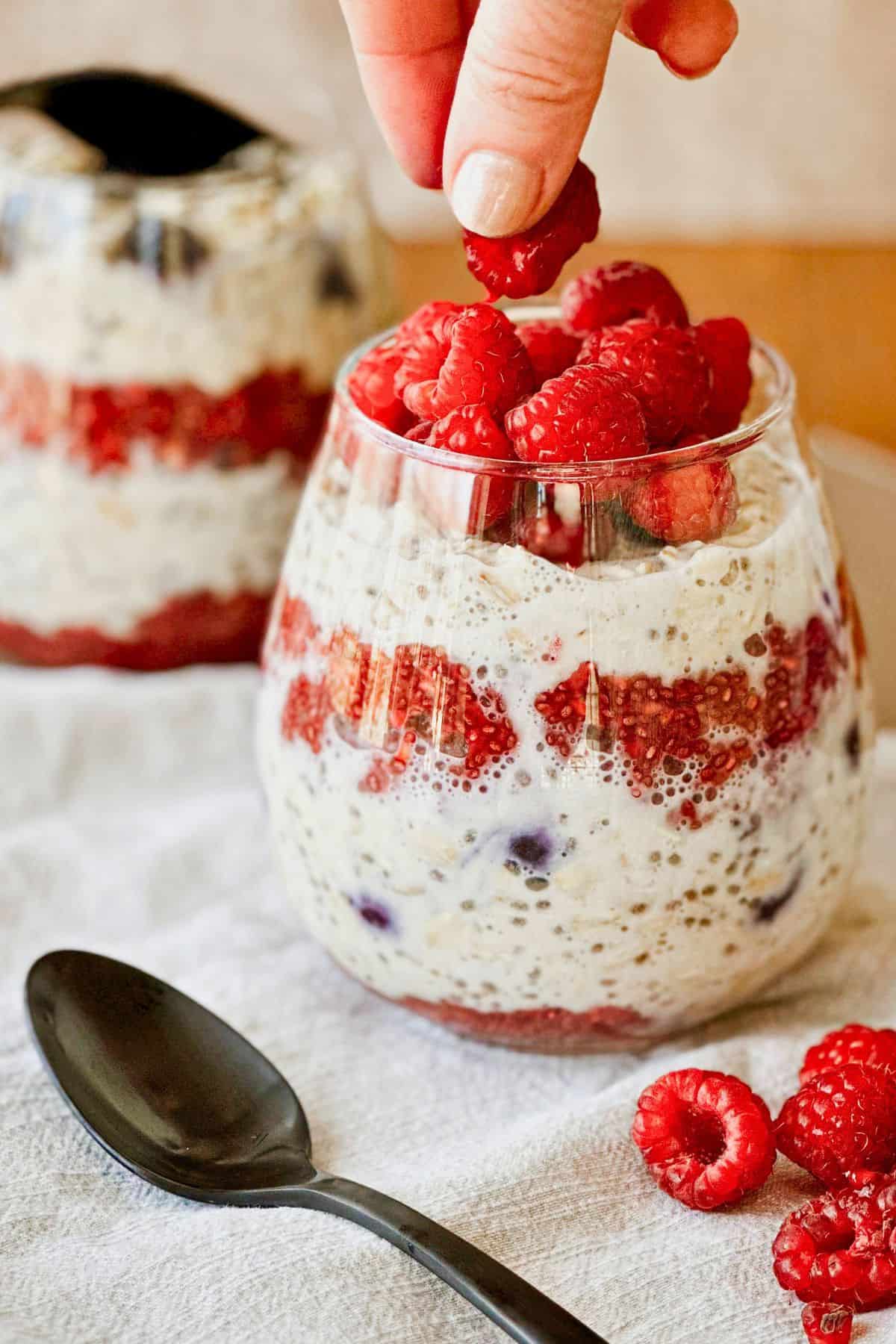 side view of Vegan Blueberry Overnight Oats with Raspberry Chia Jam in a clear glass with a hand over the top placing a raspberry on it.