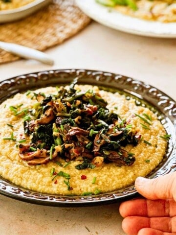 angle side view of a hand placing a plate of Vegan Grits With Savory Mushrooms and Collard Greens on a table.