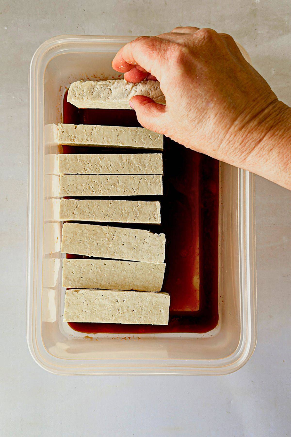 top view of hand placing tofu in a tupperware with the marinade of soy sauce and sriracha.