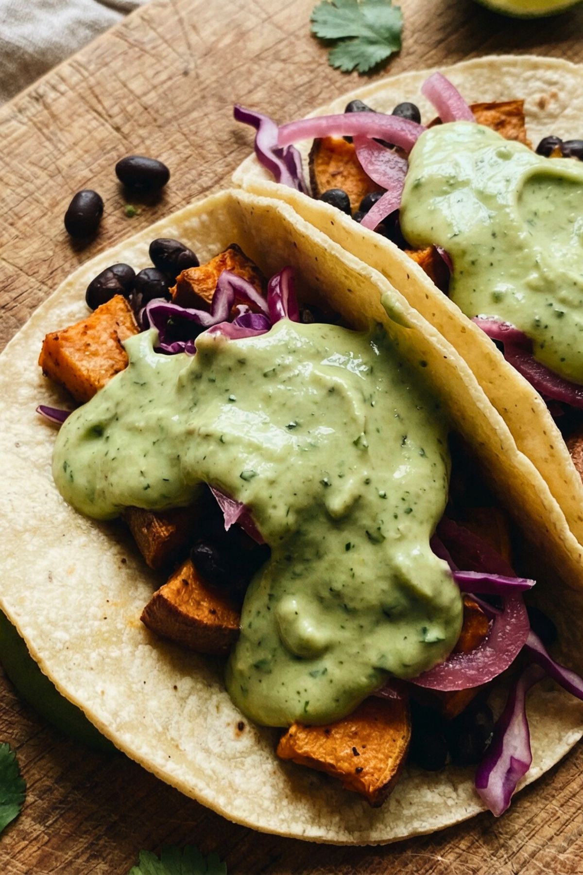 Close-up shot at a slight angle of two soft tacos on a wooden surface. The tacos are stuffed  with roasted sweet potato chunks, black beans, shredded cabbage, and pickled red onion with generous dollop of avocado crema.
