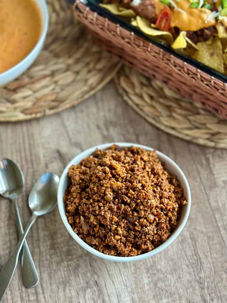 bowl of walnut meat on a table surrounded by loaded nachos, cheese sauce, and spoons