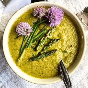 top view of creamy vegan asparagus soup in a bowl. Spoon is in the bowl. The soup is garnished with asparagus tips, chives, and chive blossoms.