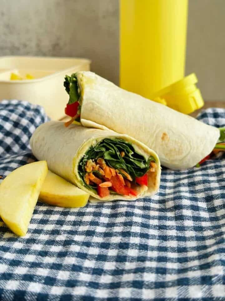 Hummus and veggie wrap on a blue checkered napkin with sliced apples next to it, yellow water bottle behind it and a bento box in the far left corner