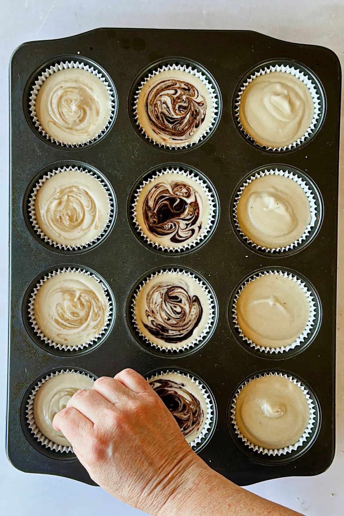 top view of hand holding a toothpick while it stirs in the peanut butter and chocolate add in to one of the cheese cakes.