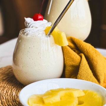 side view of a pina colada milkshake with a cherry, pineapple, and whipped cream as garnish. A plate of sliced pineapple is in front while another shake is in the back.
