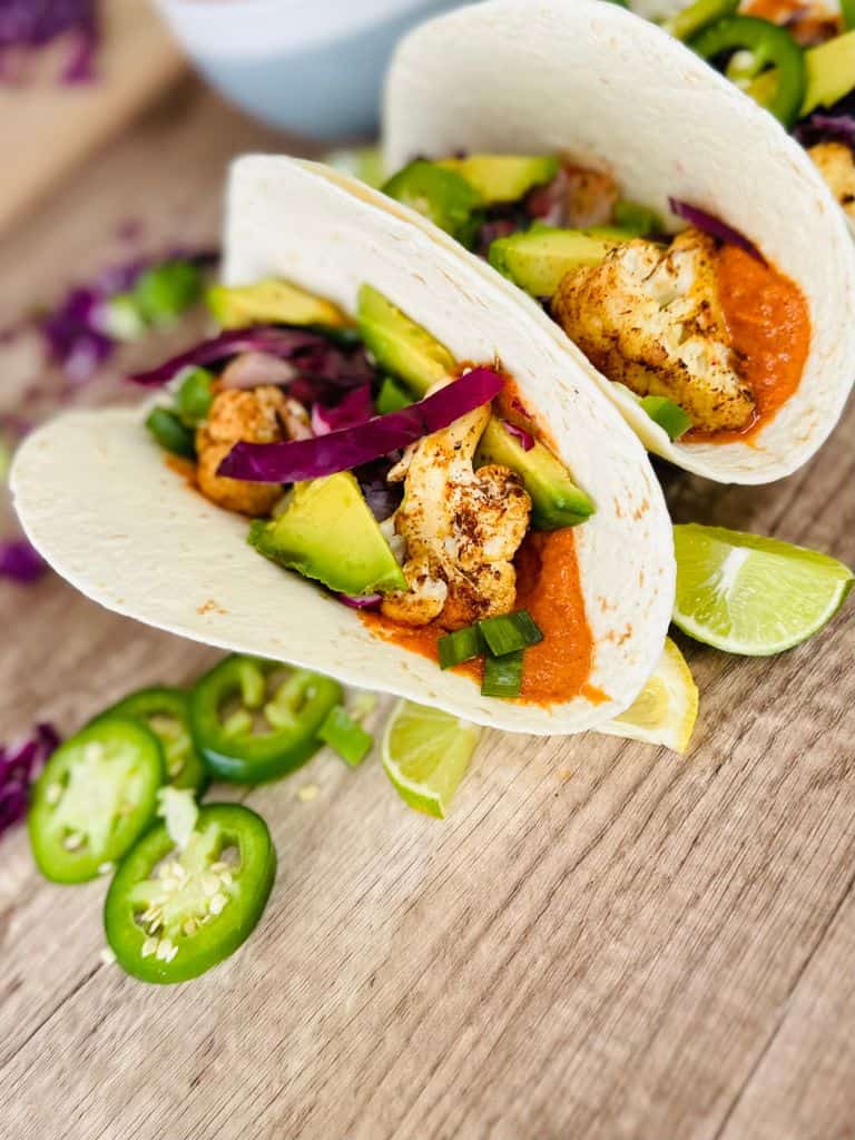 two purple cauliflower tacos with romesco sauce on wooden table surrounded by jalapeno, limes, and cabbage