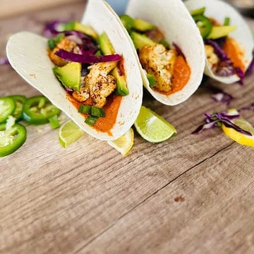set of three cauliflower tacos on table surrounded by limes, jalapeno, and purple cabbage