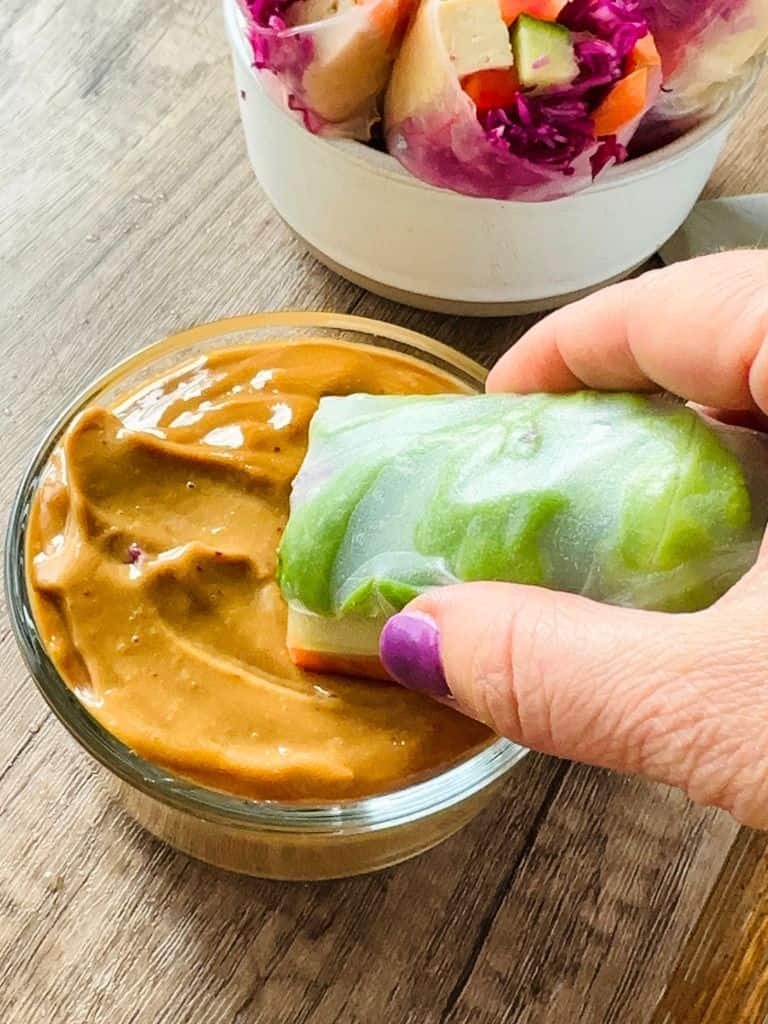 hand dipping a vegan summer roll in a bowl of peanut sauce