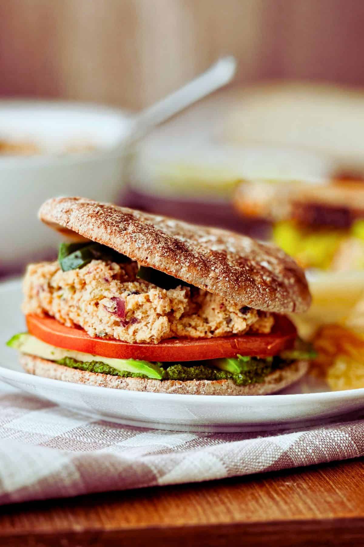 side view of tunacado sandwich; all the layers showing - pesto, avocado, tomato, vegan tuna, vegan chipotle aioli on whole wheat round bread