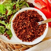 vegan bacon bit in a white bowl on a plate surrounded by a mixed salad with bacon bits and red peppers.