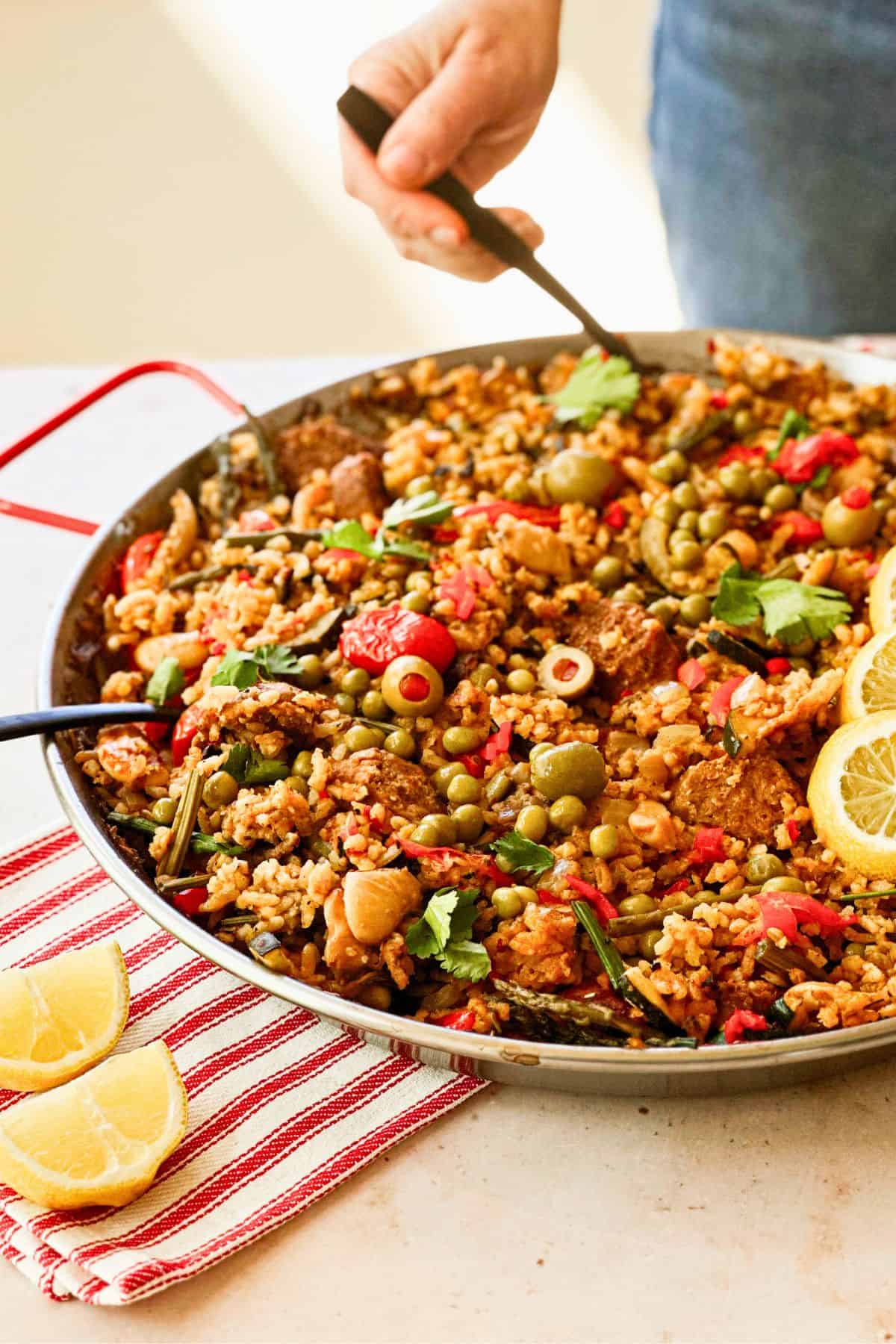 side view of vegan spanish paella in a pan with a hand in the background digging into the pan with a fork.