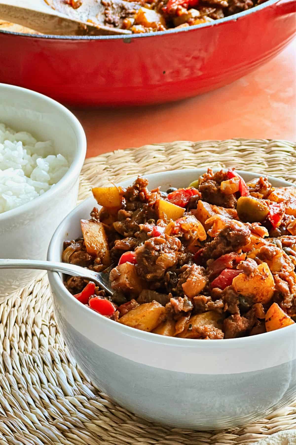 Close-up of a bowl of vegan Cuban picadillo with plant-based ground beef, potatoes, peppers, and olives in a tomato-based sauce, served with a side of white rice.