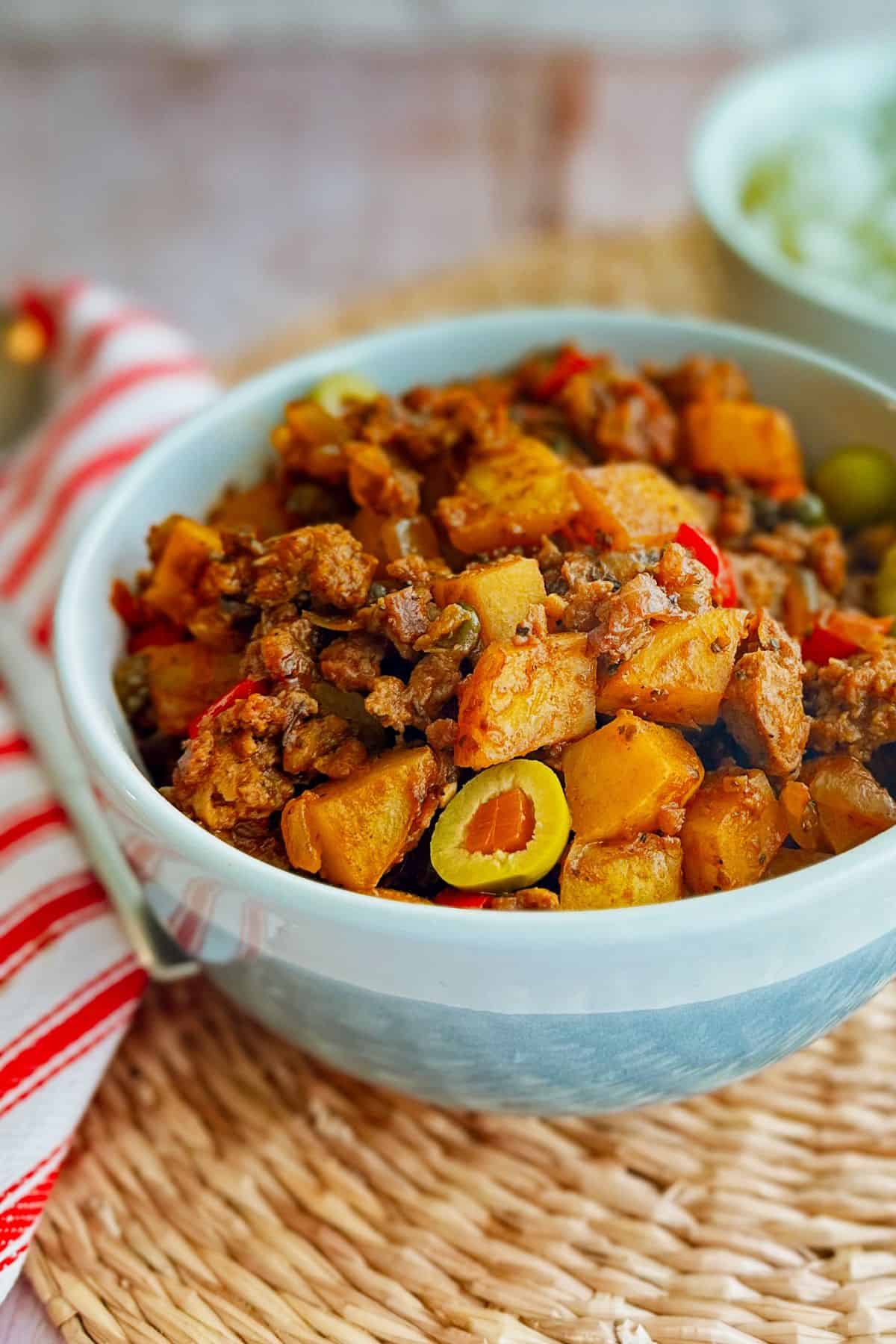 Close-up of a bowl of vegan Cuban picadillo filled with potatoes, plant-based ground beef, red peppers, onions, and green olives in a rich tomato sauce.