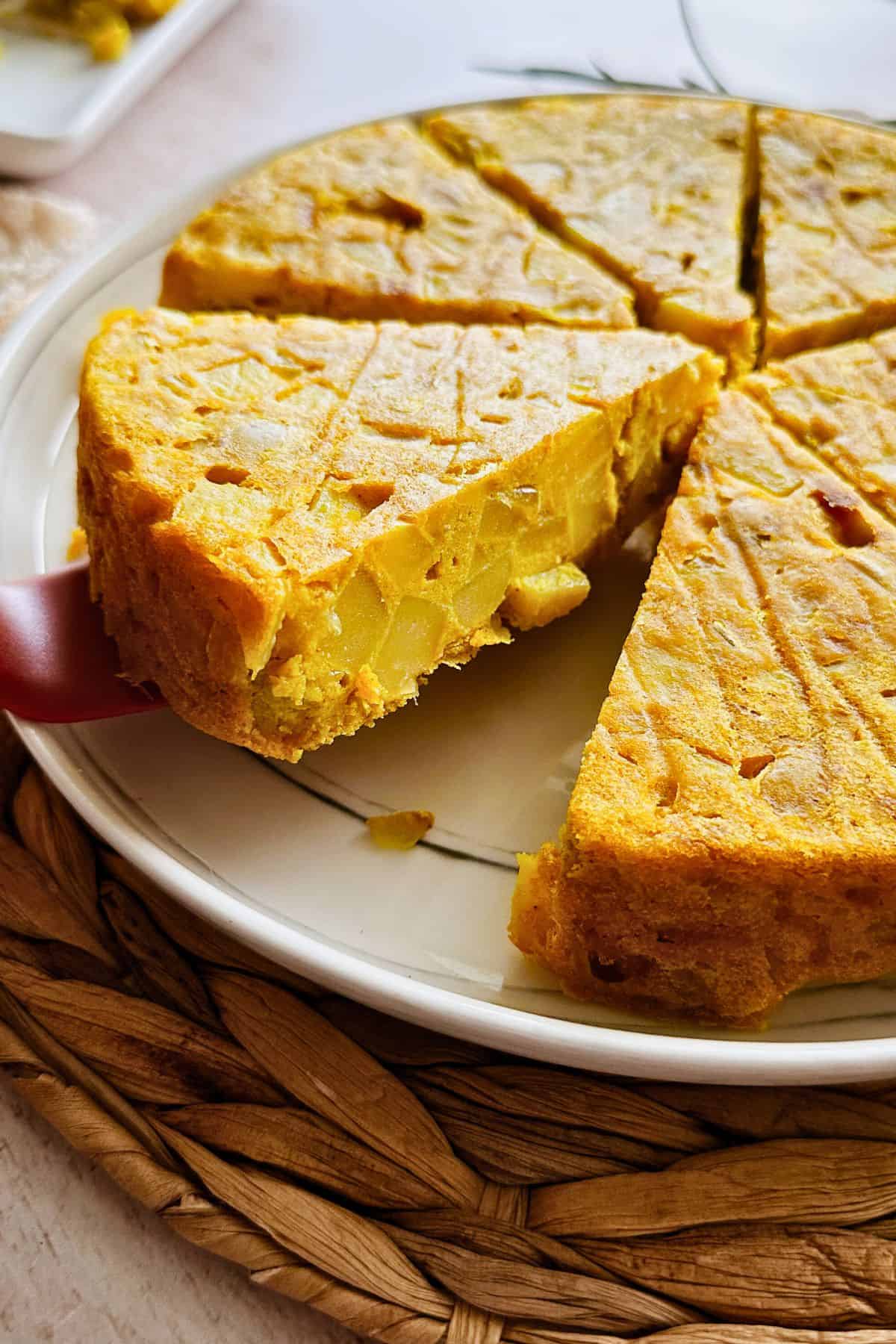 Close-up of a golden vegan Spanish tortilla (omelette) on a white plate, sliced into wedges. A spatula lifts one thick slice, showing the layered texture of potatoes and onions inside the firm, golden surface.
