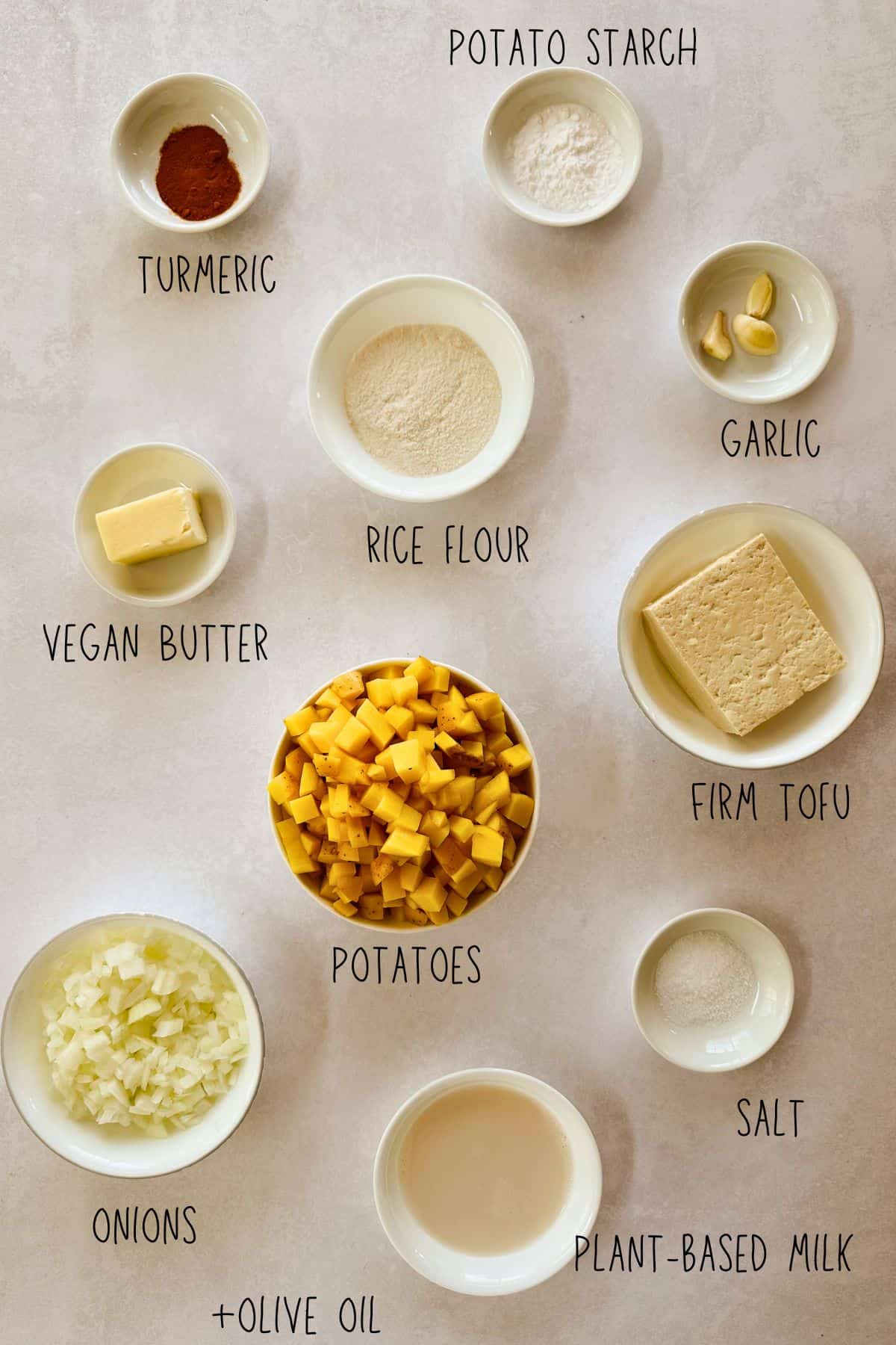 Overhead view of ingredients for a vegan Spanish tortilla (omelette) arranged in small bowls on a light surface. Labeled items include potato starch, turmeric powder, rice flour, garlic cloves, firm tofu, salt, plant-based milk, chopped onions with olive oil, diced potatoes, and vegan butter.
