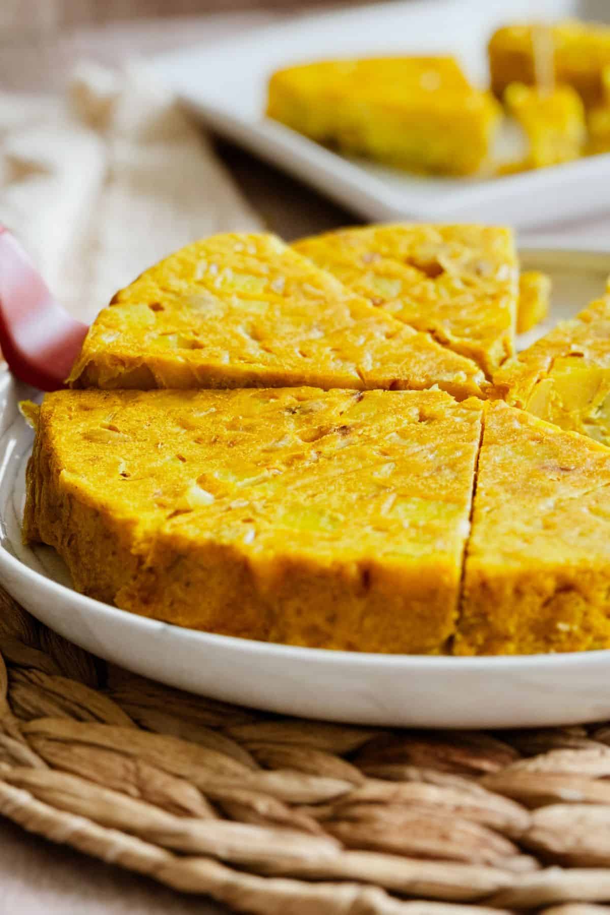 Close-up of a golden vegan Spanish tortilla (omelette) on a white plate, cut into wedges. The firm, slightly dense texture shows pieces of potato and onion inside. A serving spatula lifts one slice, with more slices visible on a plate in the background.