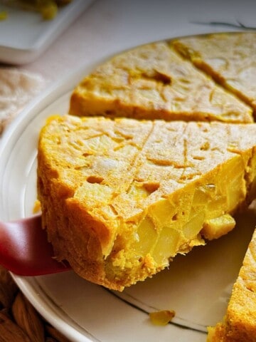 Close-up of a golden vegan Spanish tortilla (omelette) on a white plate, sliced into wedges. A spatula lifts one thick slice, showing the layered texture of potatoes and onions inside the firm, golden surface.