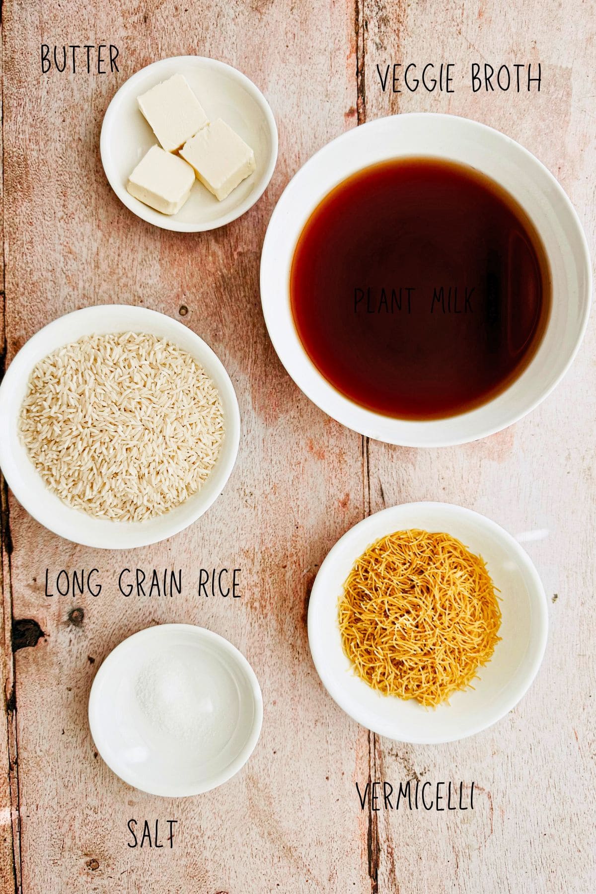 Flat lay of labeled ingredients for Arabic rice with vermicelli, including butter, veggie broth, long grain rice, vermicelli noodles, and salt, all displayed in small white bowls on a wooden surface.