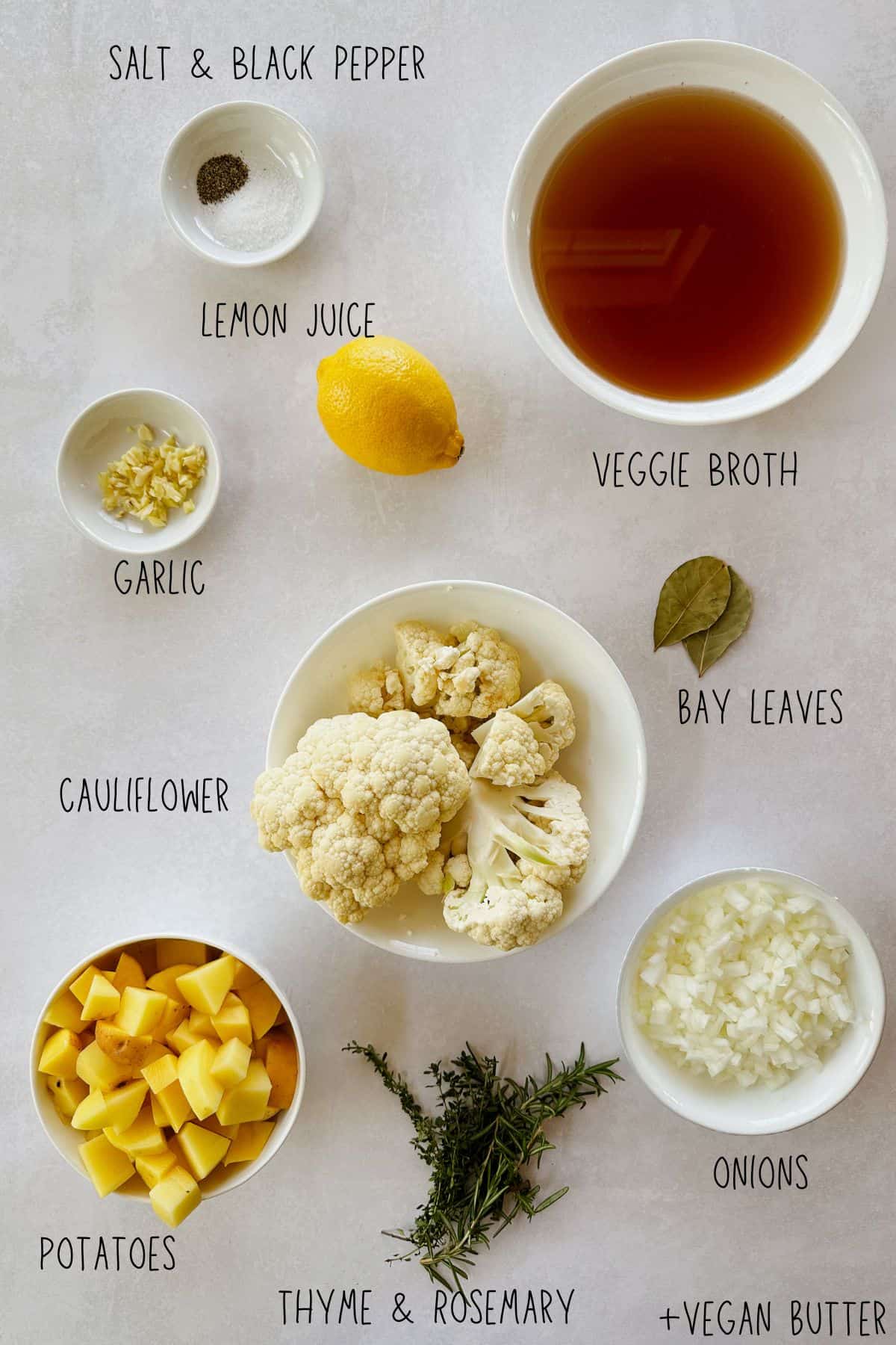 Flat lay of ingredients for vegan cauliflower potato soup including cauliflower florets, diced potatoes, onions, garlic, lemon, veggie broth, bay leaves, thyme, rosemary, salt, pepper, and vegan butter.