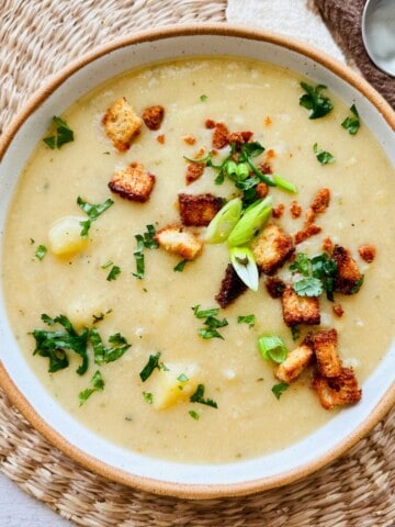 Overhead view of creamy cauliflower potato soup garnished with croutons and herbs, in a rustic bowl with a napkin and wooden spoon on the side.