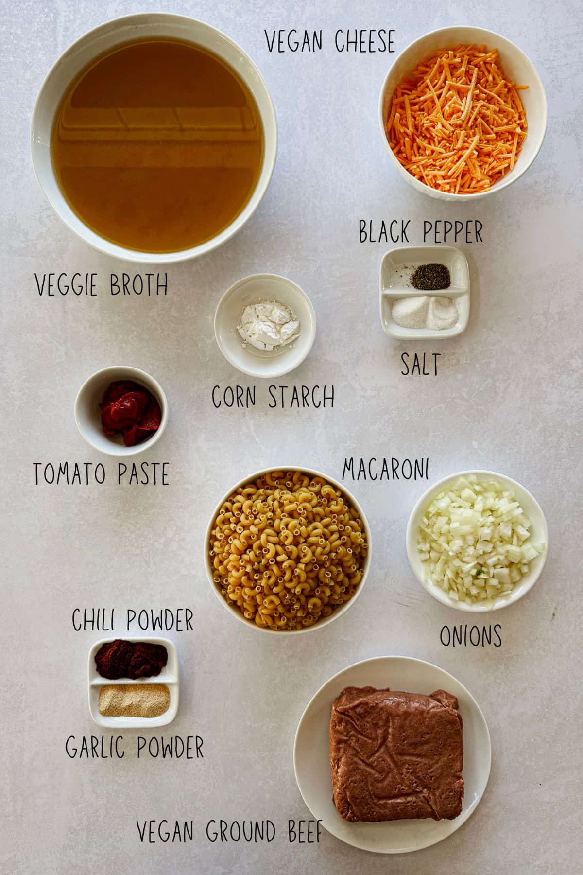 Overhead view of vegan hamburger helper ingredients arranged in bowls, including veggie broth, vegan cheese, macaroni, vegan ground beef, onion, tomato paste, cornstarch, chili powder, garlic powder, salt, and black pepper.
