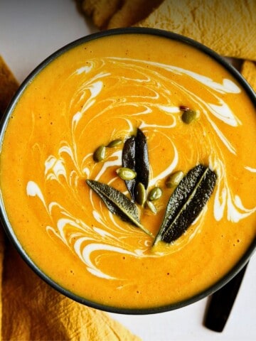 A bowl of creamy orange butternut squash and carrot soup, swirled with white dairy-free cream, topped with dark crispy sage leaves and sprinkled with green pepitas.