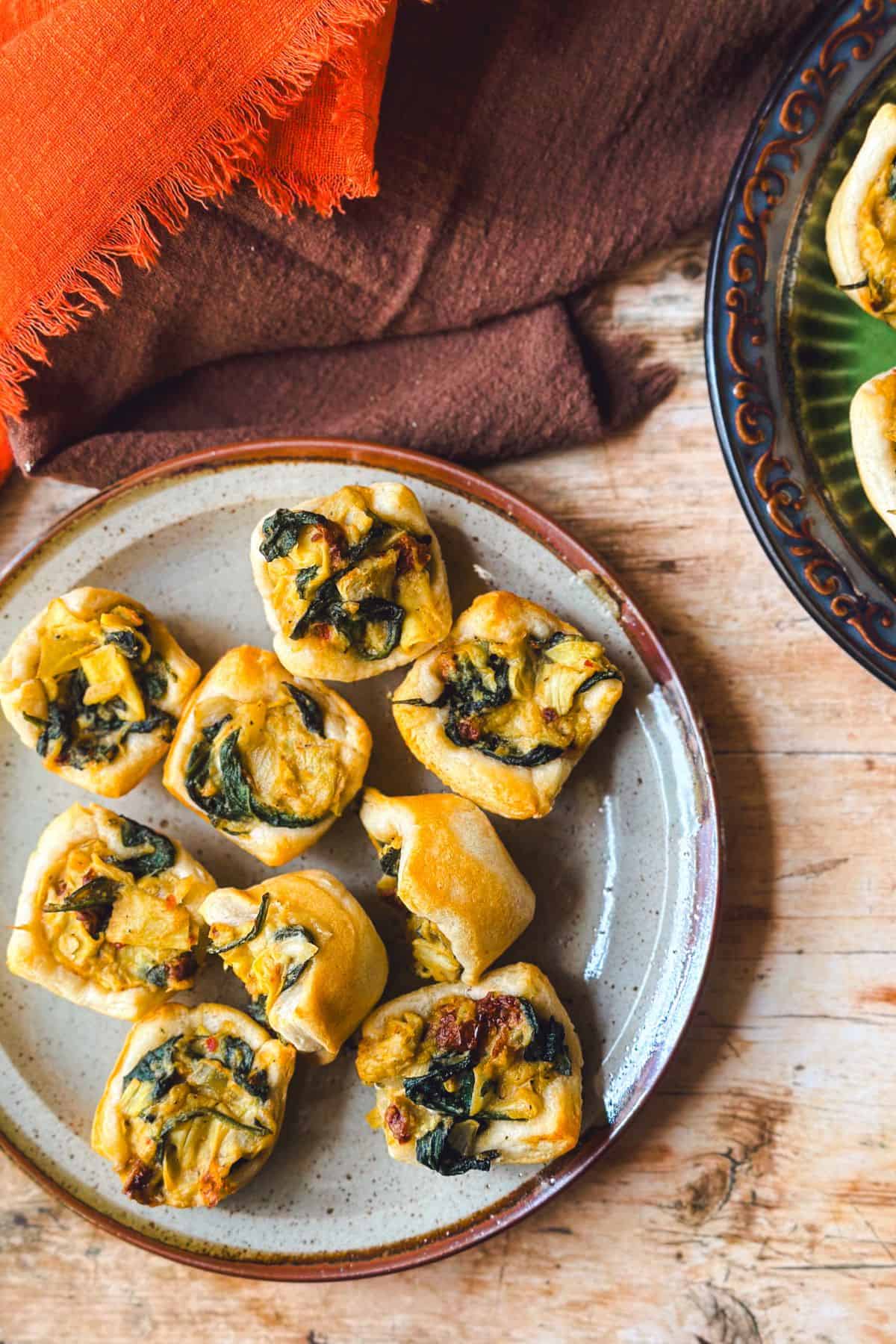 Overhead shot of golden-brown vegan spinach and artichoke bites arranged on a rustic ceramic plate, with flaky crescent roll crusts and a creamy filling made from spinach, artichokes, and sun-dried tomatoes. An orange fringed napkin and brown cloth create a cozy, autumnal backdrop.