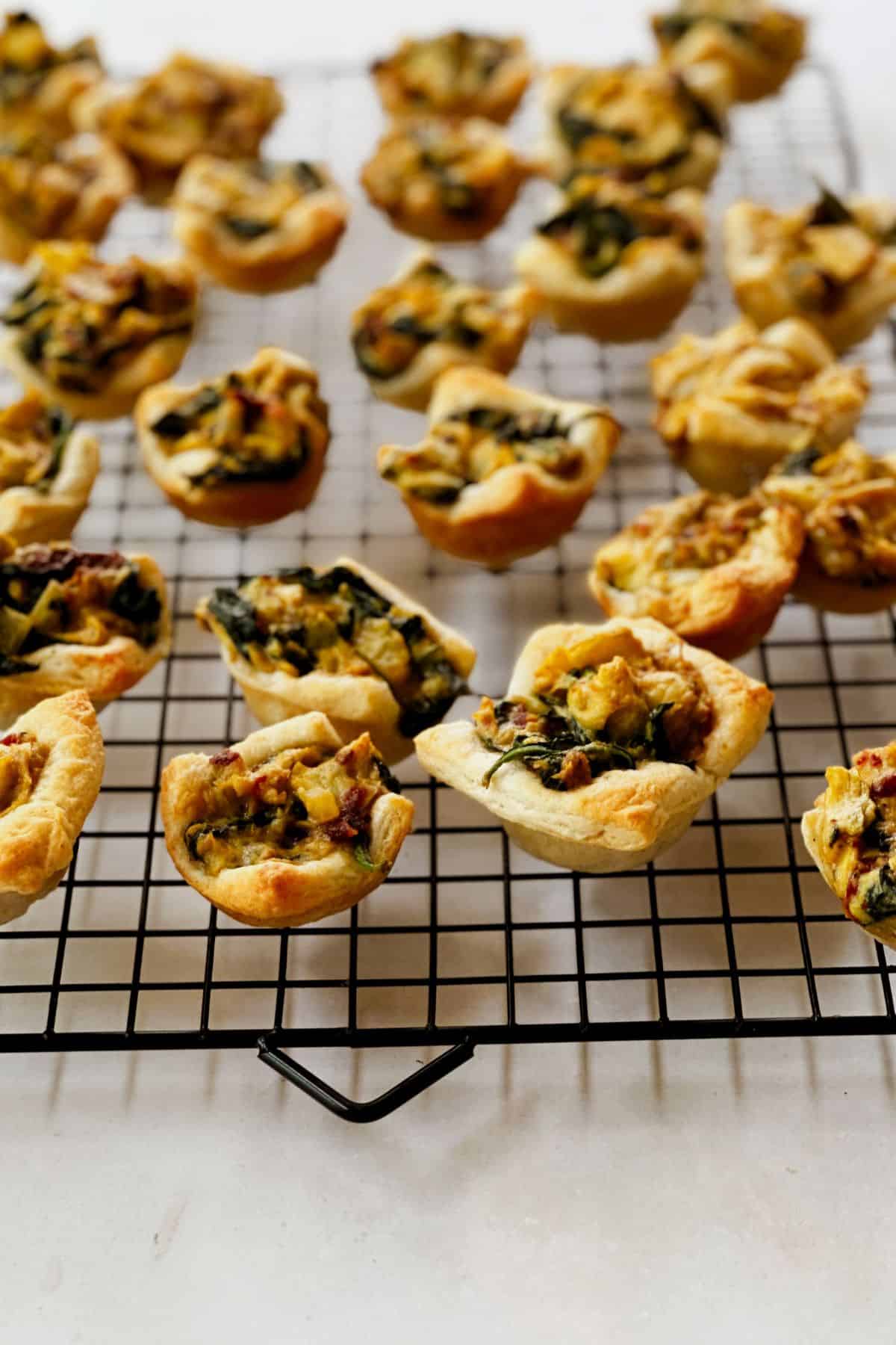 Mini vegan spinach and artichoke bites resting on a black cooling rack, showcasing golden crescent roll crusts filled with spinach, sun-dried tomatoes, and creamy filling.