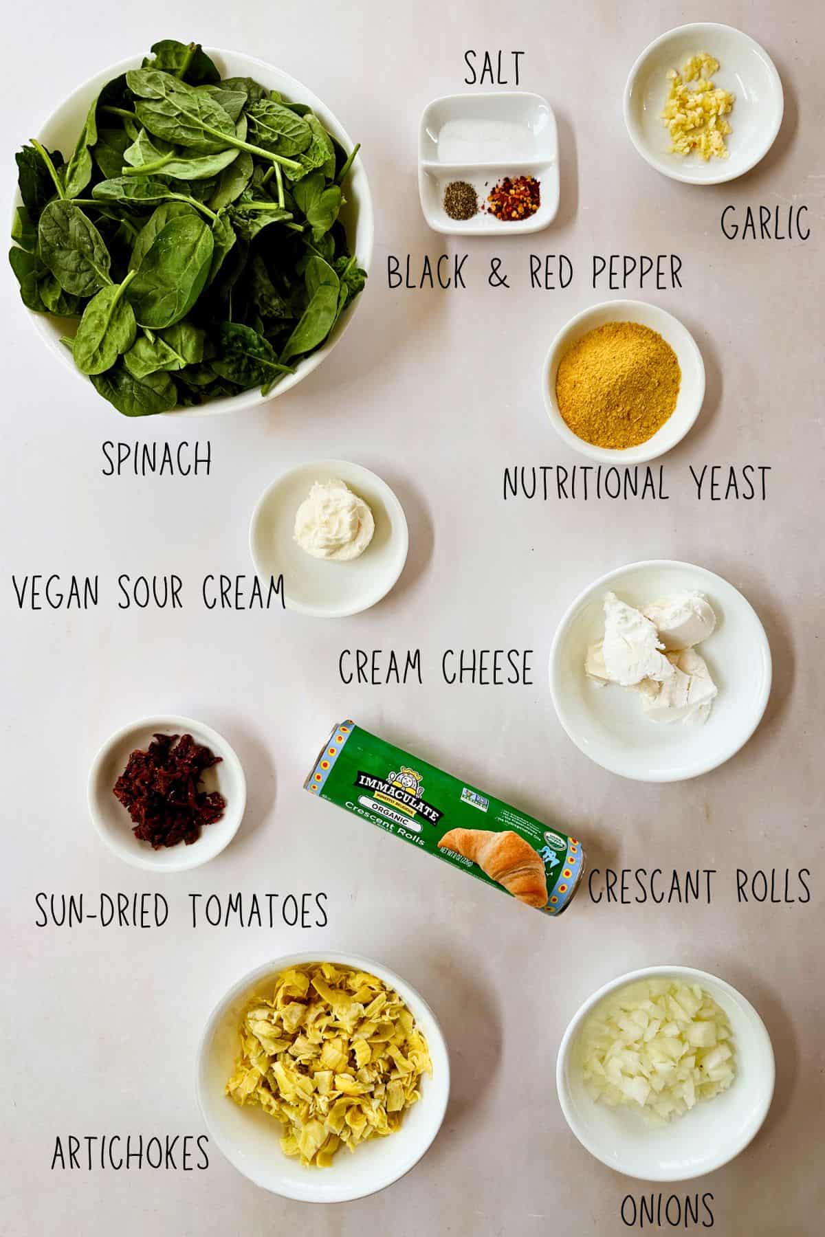 Flat lay of labeled ingredients for Spinach and Artichoke Bites, including bowls of spinach, artichokes, diced onions, garlic, sun-dried tomatoes, nutritional yeast, vegan sour cream, cream cheese, spices, and a tube of crescent rolls on a light surface.