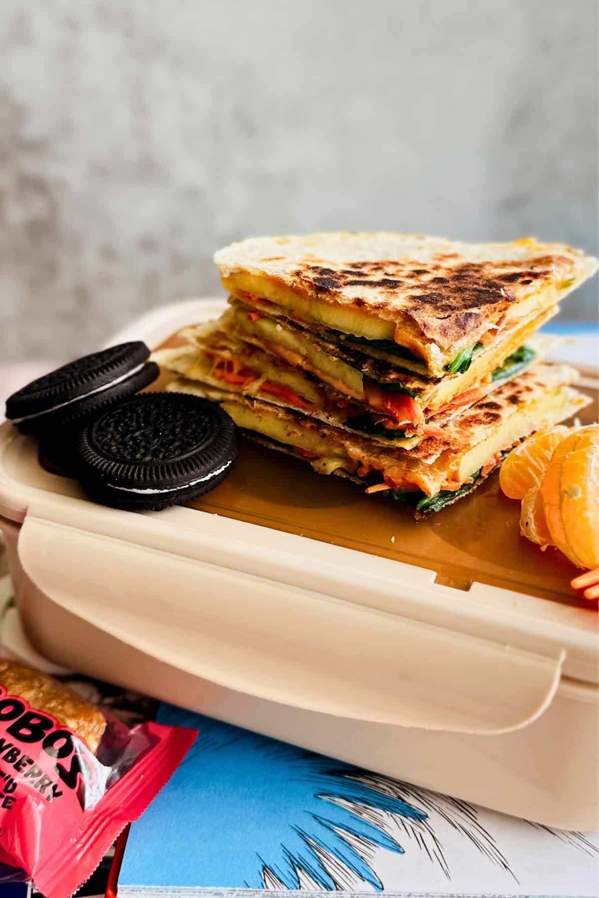 A close-up of stacked apple and veggie quesadilla wedges on top of a beige lunchbox, served with two chocolate sandwich cookies and a peeled mandarin orange. A colorful snack bar wrapper and a blue illustrated book page are partially visible underneath.