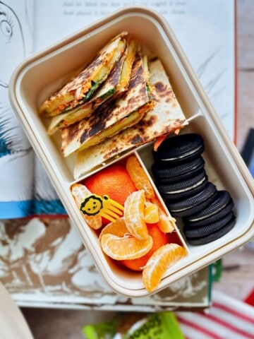 A lunchbox with sliced apple and veggie quesadillas stacked in one compartment, paired with mandarin orange segments and chocolate sandwich cookies in another side compartments. A small yellow lion-shaped food pick is placed among the orange slices. A Dr. Seuss book and snack packaging are visible in the background.