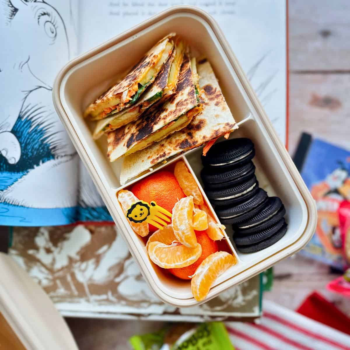 A lunchbox with sliced apple and veggie quesadillas stacked in one compartment, paired with mandarin orange segments and chocolate sandwich cookies in another side compartments. A small yellow lion-shaped food pick is placed among the orange slices. A Dr. Seuss book and snack packaging are visible in the background.