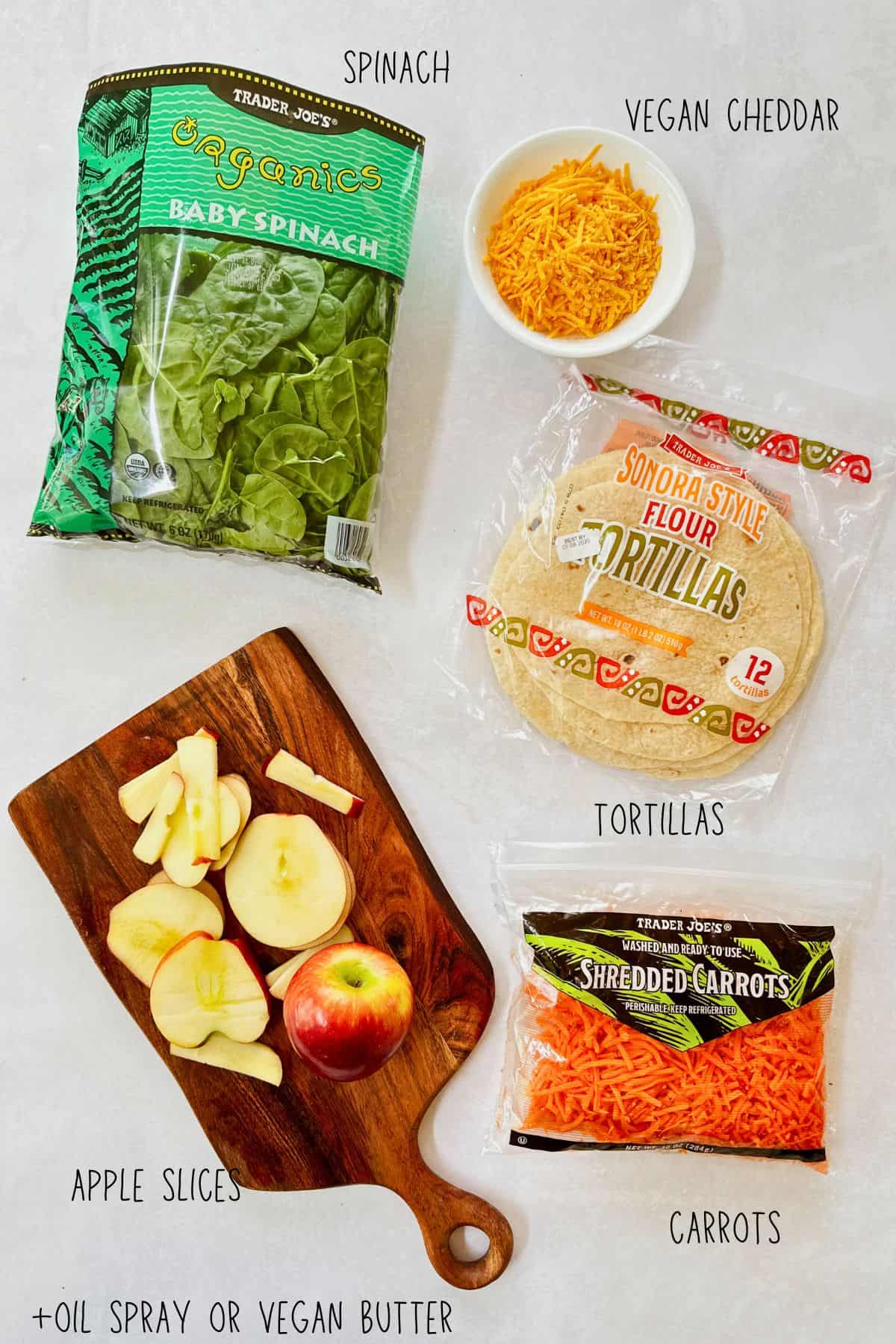 Flat lay of ingredients for Apple and Veggie Quesadillas including baby spinach, shredded vegan cheddar, flour tortillas, shredded carrots, and sliced apples on a small wooden cutting board, plus a note for optional oil spray or vegan butter. All ingredients are labeled.